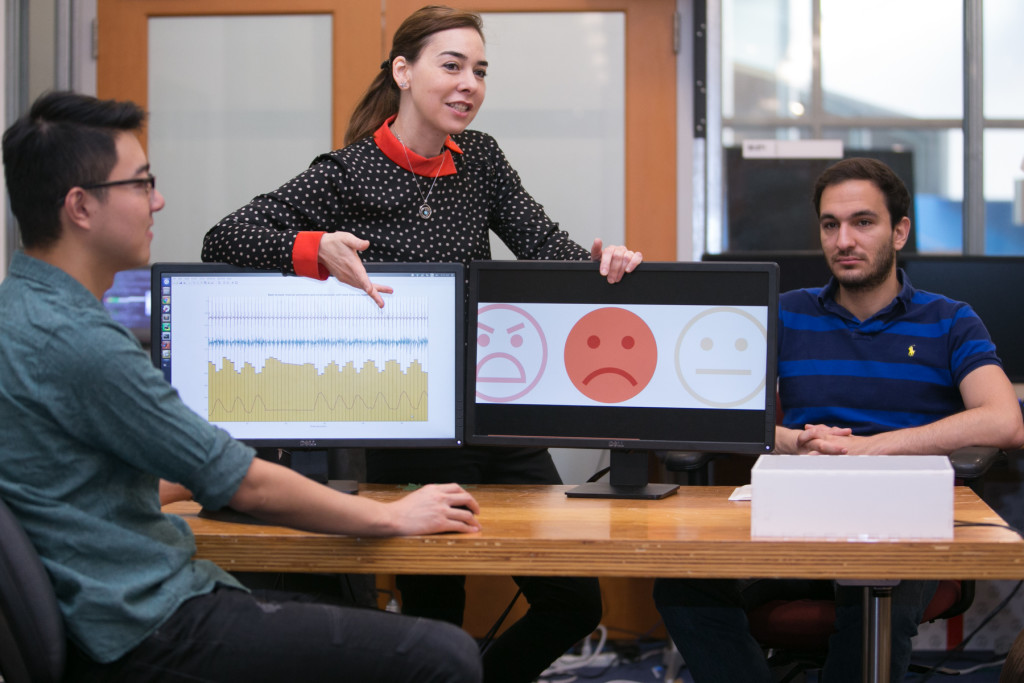 Professor Dina Katabi (middle) explains how PhD Fadel Adib's face (right) is neutral, but that EQ-Radio's analysis of his heartbeat and breathing show that he is sad. Credit: Jason Dorfman MIT CSAIL
