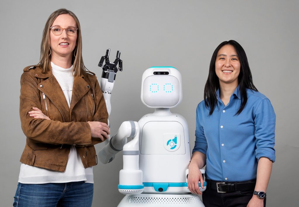 Two women stand next to a friendly-looking humanoid robot with one arm.