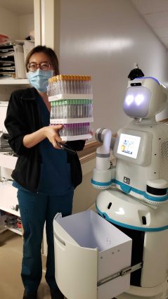 A smiling clinical staff member holds a large stack of samples. Next to her, a humanoid robot almost as tall as she is holds its storage container open. The storage container has plenty of room for this large stack of samples. 